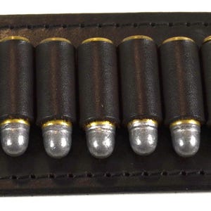 Könnte beinhalten: Ein dunkelbrauner Leder-Patronengürtel mit 13 dunkelbraunen Patronen mit silbernen Spitzen und Messinghülsen. Der Gürtel hat zwei hellbraune Lederschlaufen und ist für das Tragen über der Schulter konzipiert.