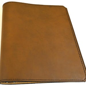 Può includere: Una copertina rettangolare marrone per taccuino con una finitura liscia e opaca. La copertina presenta cuciture marrone scuro lungo tutto il perimetro, con un dorso arrotondato sul lato sinistro e un angolo superiore destro leggermente inclinato.