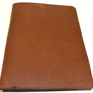 May include: A brown leather journal with a stitched border. The cover is a solid, warm brown colour with a smooth texture. The edges are neatly stitched with a contrasting thread, adding a touch of detail to the design. The journal appears to be closed.