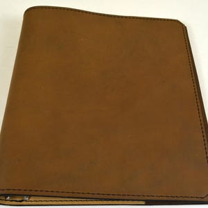May include: A rectangular brown leather binder featuring a smooth matte finish and dark brown stitching along the edges. The binder has a rounded spine on the left side and a single angled corner on the top right, with visible metal ring hardware.