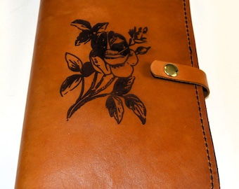 May include: A brown leather journal with a black rose design embossed on the front cover. The journal has a brown leather strap with a gold metal buckle.