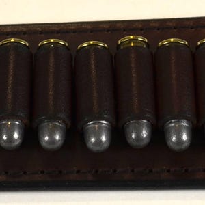 Può includere: Un portacartucce in pelle marrone scuro di forma rettangolare con estremità arrotondate e bordi cuciti. Contiene dodici scomparti individuali, ognuno dei quali ospita un proiettile metallico con bossolo in ottone e punta in piombo argentato.
