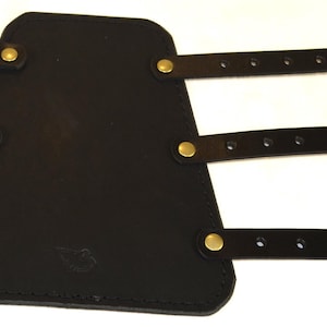 May include: Black leather arm guard with three adjustable straps and brass buckles. The guard has a triangular shape with rounded edges and is secured with three straps.