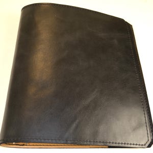 May include: A black leather notebook cover with a smooth, matte finish and visible stitching along the edges. The cover features a rounded spine on the left side and a straight, angled edge on the right side, revealing a light tan interior lining.