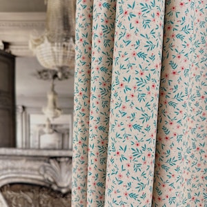 May include: Close-up of a cream-coloured fabric curtain with a repeating pattern of tiny pink flowers and teal leaves. The fabric is draped in soft folds. The floral design creates a delicate, natural aesthetic, suitable for home decor.
