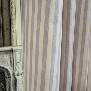 May include: Close-up of a fabric with vertical stripes in pale lavender and cream. The striped pattern is visible on the curtain fabric. The fabric is likely used for home decor, such as curtains or upholstery.