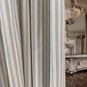May include: Close-up of a fabric curtain with vertical stripes. The stripes alternate between a light blue and a cream color. The curtain is slightly draped, showing the texture of the fabric.