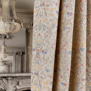 May include: Close-up of beige floral patterned curtains. The fabric features a repeating pattern of small flowers in shades of yellow, pink, and blue, set against a neutral background. The curtains are partially drawn, revealing a glimpse of a chandelier and a room.