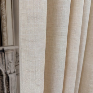 May include: Close-up of beige fabric curtains with a subtle plaid pattern. The curtains are folded in soft, vertical pleats, creating a textured and elegant look. The neutral color and simple design suggest a classic, versatile style suitable for various interior settings.