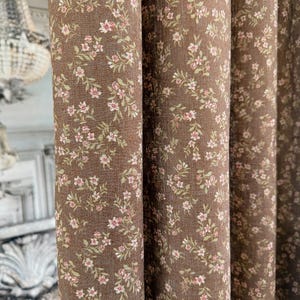 May include: Close-up of brown floral curtains. The fabric features a repeating pattern of small pink flowers and green leaves. The curtains are made of a textured material and are partially draped, showing the folds and depth of the fabric.