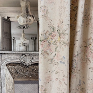 May include: A beige curtain featuring a repeating floral pattern with soft pink roses, green leaves, and small blue flowers. The fabric has a textured appearance and hangs in vertical folds, partially obscuring a room with a fireplace and chandelier.