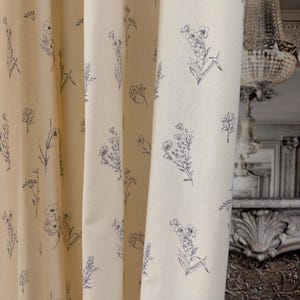 May include: Cream-colored fabric with a repeating pattern of delicate, dark blue floral illustrations. The fabric is draped in soft folds, showcasing the detailed botanical designs. The overall aesthetic is a blend of rustic charm and elegant simplicity, suitable for home decor.