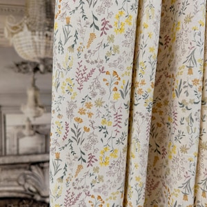 May include: A cream-coloured fabric curtain featuring a repeating pattern of small wildflowers in shades of yellow, muted green, and soft pink. The textile displays a fine weave texture and hangs in vertical folds, showcasing the delicate floral print.