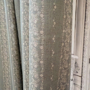 May include: Close-up of a pale blue curtain with a floral design. The curtain features vertical stripes of small white flowers and leaves. The fabric appears to be linen or a similar textured material. The curtain is partially folded.