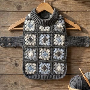 Puede incluir: Un suéter de ganchillo para mascota con un patrón de cuadrados de la abuela en tonos gris, blanco y azul claro. La prenda tiene un cuello alto acanalado gris oscuro, bordes acanalados a juego y dos correas laterales con broches de presión.