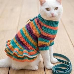 May include: A white cat wearing a crocheted sweater with horizontal stripes in teal, orange, and purple. The sweater features a ruffled hem, a ribbed collar, and a gold-toned metal D-ring attachment on the back, accompanied by a matching teal leash.