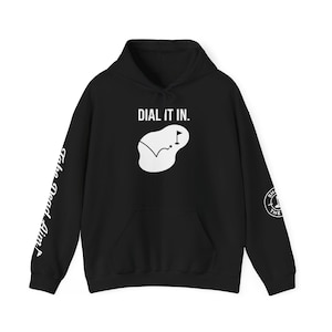 Puede incluir: Sudadera con capucha negra con el texto "DIAL IT IN." sobre un gráfico blanco de campo de golf. La manga izquierda tiene el texto "Take Dead Aim" y la manga derecha un logotipo circular.