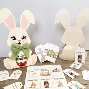 May include: Two white Easter bunny cutouts with pink inner ears and a green bow. One bunny holds a Kinder egg. Several gift tags with Easter-themed graphics and text, including "Happy Easter!" and "Some-bunny loves you!"