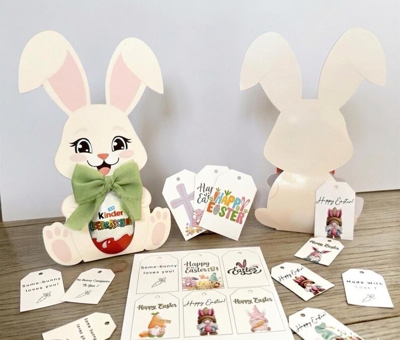 May include: Two white Easter bunny cutouts with pink inner ears and a green bow. One bunny holds a Kinder egg. Several gift tags with Easter-themed graphics and text, including "Happy Easter!" and "Some-bunny loves you!"
