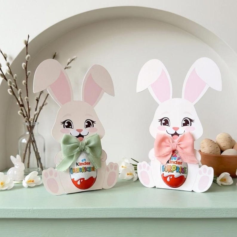 Two cute paper Easter bunny Kinder egg holders in pink and white, wearing green and pink fabric bows, standing on a light green surface with decorative willow branches and mini Easter eggs in the background