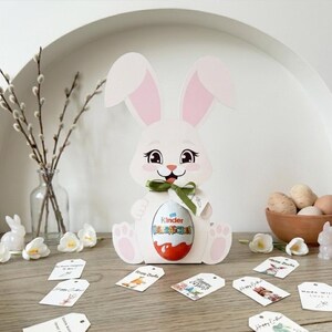 May include: A white Easter bunny-shaped gift box holding a Kinder Surprise egg, with pink inner ears and a green ribbon. The scene includes Easter eggs in a wooden bowl, decorative tags, and floral accents, creating a festive holiday display.