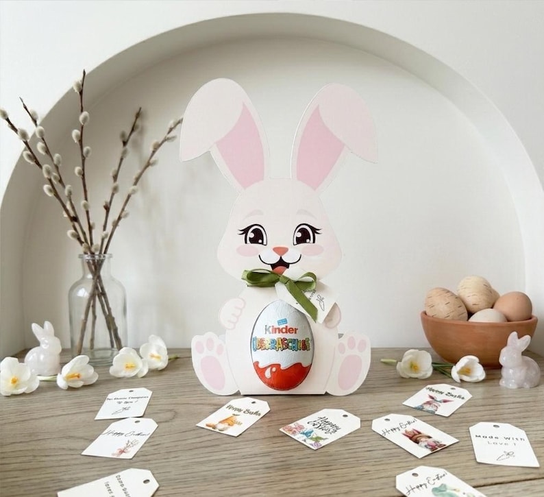 May include: A white Easter bunny-shaped gift box holding a Kinder Surprise egg, with pink inner ears and a green ribbon. The scene includes Easter eggs in a wooden bowl, decorative tags, and floral accents, creating a festive holiday display.