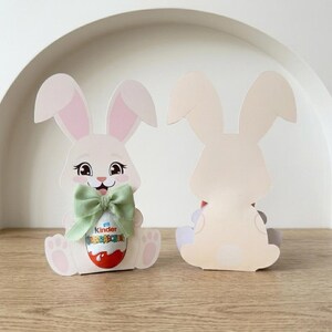 May include: Two Easter bunny-shaped paper cutouts. The front bunny has a green bow and holds a Kinder Surprise egg. The bunny is white with pink inner ears and a smiling face. The second bunny is a plain cutout of the bunny's back.