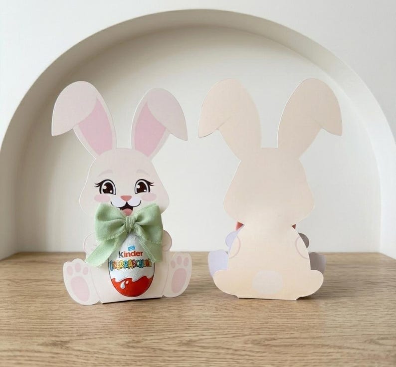 May include: Two Easter bunny-shaped paper cutouts. The front bunny has a green bow and holds a Kinder Surprise egg. The bunny is white with pink inner ears and a smiling face. The second bunny is a plain cutout of the bunny's back.