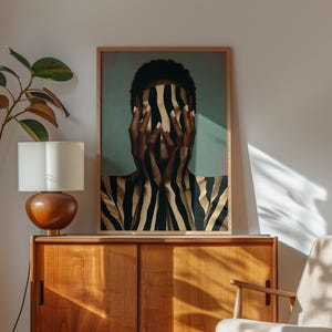 May include: A framed rectangular wall art print featuring a person with dark skin covering their face with hands, both adorned with vertical black and gold stripes. The figure is set against a muted teal background, presented in a light wood frame.