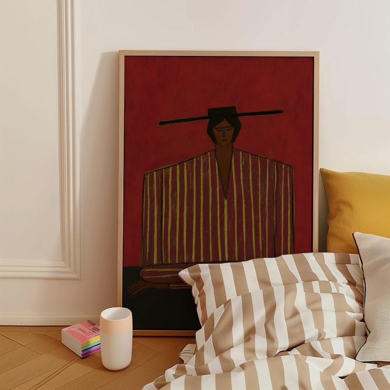 May include: A framed wall art print featuring a stylized figure wearing a wide-brimmed hat and a vertically striped garment in shades of yellow and brown, set against a deep red background. The artwork is displayed in a light wood frame.