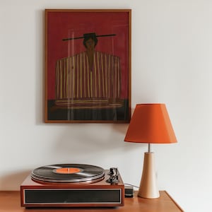 May include: A framed wall art print featuring a stylized figure in a striped garment against a deep red background, positioned above a wooden record player and a table lamp with a vibrant orange lampshade and a light beige conical base.