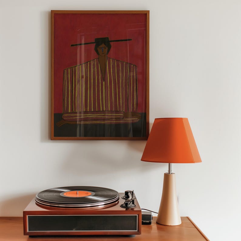 May include: A framed wall art print featuring a stylized figure in a striped garment against a deep red background, positioned above a wooden record player and a table lamp with a vibrant orange lampshade and a light beige conical base.