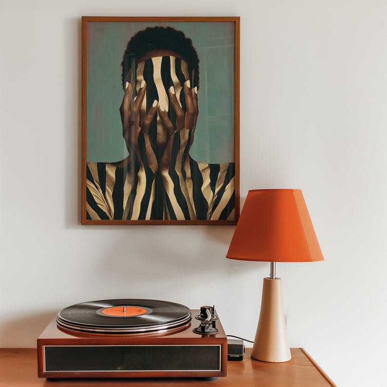 May include: A framed wall art print featuring a person with hands covering their face, displaying a black and gold zebra-striped pattern. Beside it sits a wooden record player with a black vinyl disc and a lamp with a bright orange conical shade.