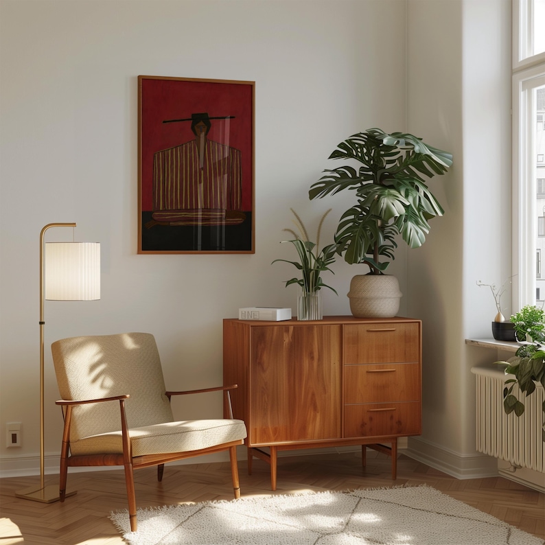 May include: A room interior featuring a beige upholstered armchair with wooden armrests, a wooden cabinet with three drawers and a door, a tall floor lamp with a white pleated shade, and a framed wall art piece depicting a figure in a hat.