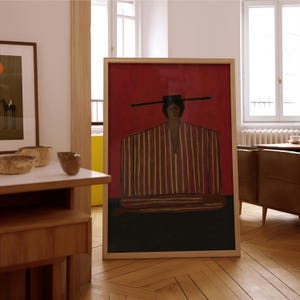 May include: A framed rectangular wall art print featuring a stylized figure wearing a wide-brimmed hat and a long-sleeved garment with vertical yellow and red stripes. The figure is set against a deep red background with a dark brown base.