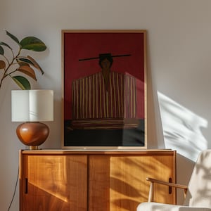May include: A framed rectangular wall art print featuring a stylized figure wearing a wide-brimmed hat and a garment with vertical red and yellow stripes against a deep red background, set within a light-colored wooden frame on a flat surface.