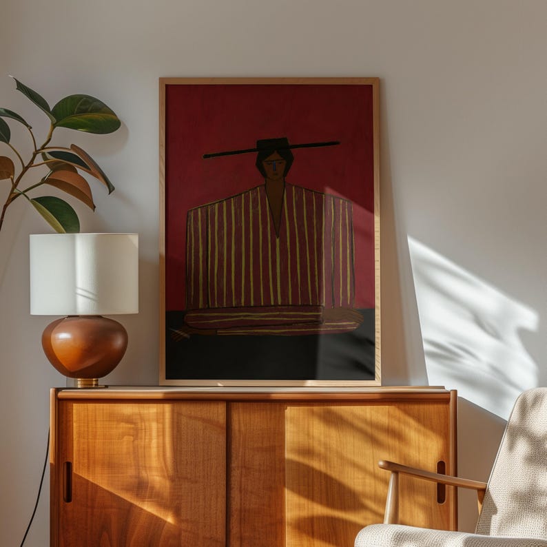 May include: A framed rectangular wall art print featuring a stylized figure wearing a wide-brimmed hat and a garment with vertical red and yellow stripes against a deep red background, set within a light-colored wooden frame on a flat surface.