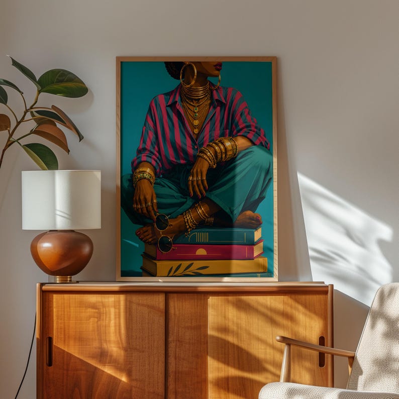 May include: A framed wall art print depicting a person wearing a pink and blue striped shirt and teal trousers, adorned with gold hoop earrings, layered necklaces, and multiple gold bracelets, sitting cross-legged on a stack of three colorful books.