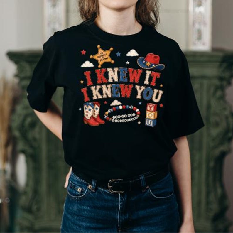 K&ouml;nnte beinhalten: Ein schwarzes T-Shirt mit einem Grafikdruck und dem Text I KNEW IT I KNEW YOU. Das Design zeigt einen Sheriffstern, einen Cowboyhut, Cowboystiefel, ein Perlenarmband und gestapelte Bl&ouml;cke mit der Aufschrift YOU in Rot, Wei&szlig; und Blau.