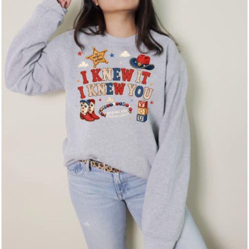 K&ouml;nnte beinhalten: Ein hellgraues Sweatshirt mit Rundhalsausschnitt und einem Grafikdruck mit dem Text I KNEW IT I KNEW YOU. Das Design zeigt einen gelben Sheriffstern, einen rot-blauen Cowboyhut, rot-wei&szlig;e Cowboystiefel, ein Perlenarmband und gestapelte Bl&ouml;cke.