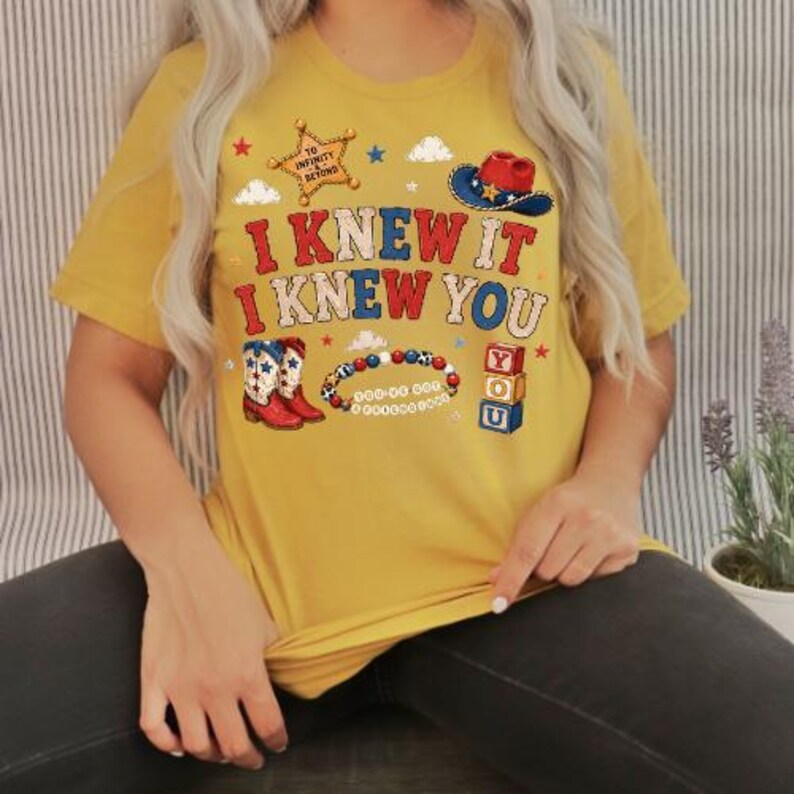 K&ouml;nnte beinhalten: Ein senfgelbes T-Shirt mit einem Grafikdruck mit dem Text I KNEW IT I KNEW YOU, einem Sheriffstern mit TO INFINITY AND BEYOND, einem Cowboyhut, einem Paar Westernstiefeln, einem Perlenarmband und drei gestapelten Buchstabenw&uuml;rfeln.