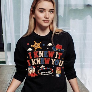 K&ouml;nnte beinhalten: Ein schwarzes Langarm-Sweatshirt mit einem Grafikdruck mit dem Text I KNEW IT I KNEW YOU, einem gelben Sheriffstern mit TO INFINITY AND BEYOND, einem Cowboyhut, rot-wei&szlig;en Stiefeln, einem Perlenarmband und gestapelten Bl&ouml;cken mit den Buchstaben Y O U.