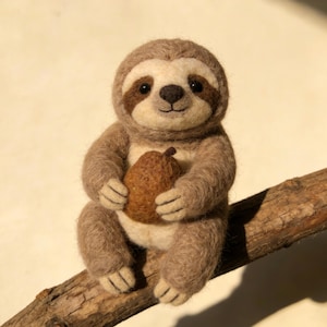 May include: A handmade felt sloth figurine, light brown with a cream-colored face and dark brown accents. The sloth is holding a brown pear-shaped object and is perched on a wooden branch. The sloth has a smiling expression.