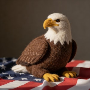 May include: A handcrafted felt bald eagle figurine with a white head, brown body, and yellow beak and feet. The eagle is posed on a red, white, and blue American flag.