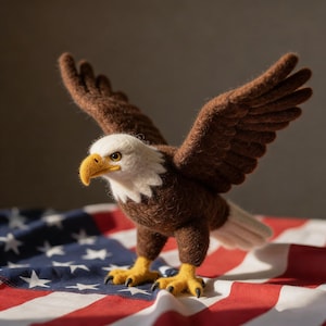 May include: A handcrafted felt eagle with outstretched wings, featuring brown feathers, a white head, and a yellow beak and talons. The eagle stands on a red, white, and blue American flag, showcasing patriotic colors and themes.