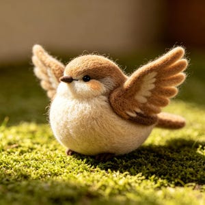 Puede incluir: Un pájaro de fieltro hecho a mano con las alas extendidas. El pájaro tiene la cabeza y las alas marrones, el cuerpo color crema y un pico pequeño y oscuro. Está posado sobre musgo verde, creando una escena natural y caprichosa.