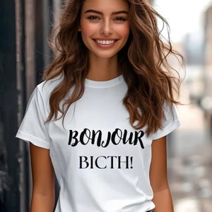 Puede incluir: Camiseta blanca con las palabras "BONJOUR BICTH!" en escritura negra. La camiseta es de manga corta y tiene cuello redondo. La imagen muestra a una persona usando la camiseta.