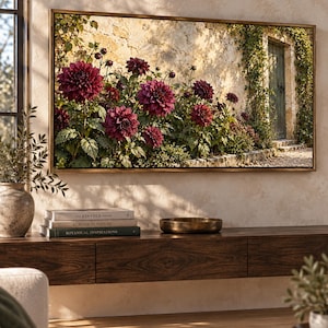 May include: A rectangular wall art piece in a thin bronze frame displaying a landscape photograph of deep burgundy dahlia flowers blooming in front of a weathered stone wall with a wooden door, mounted above a dark wood floating console table.