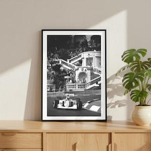 May include: Black and white framed print of a Formula 1 race car speeding around a track. The image features a historic race scene with a grandstand and lush greenery in the background. The car has the word "Marlboro" on it.
