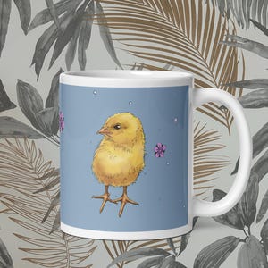Könnte beinhalten: Weiße Keramiktasse mit einer Illustration eines gelben Küken auf hellblauem Hintergrund. Das Küken hat orangefarbene Füße und einen gelben Körper. Rosa Blumen und kleine weiße Punkte sind um das Küken herum verstreut. Die Tasse hat einen weißen Henkel.